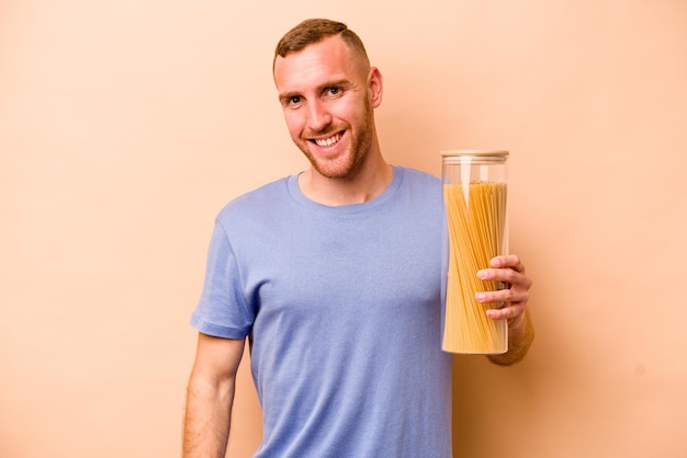 Joven caucásico sosteniendo tarro de espaguetis aislado sobre fondo beige feliz sonriente y alegre