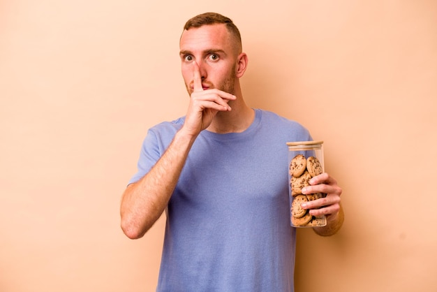 Joven caucásico sosteniendo un tarro de galletas aislado de fondo beige guardando un secreto o pidiendo silencio