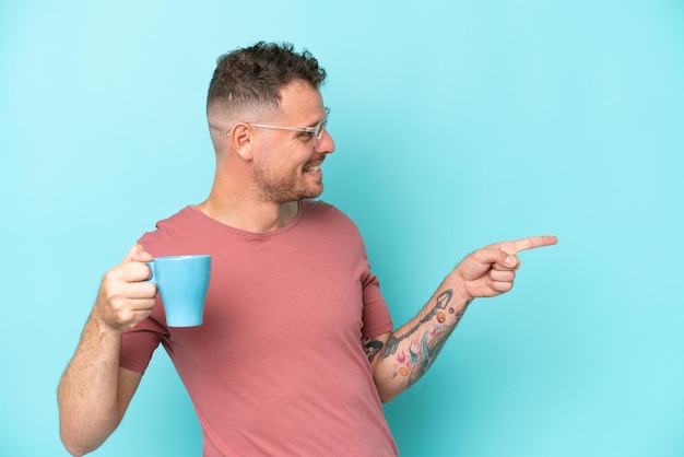 Joven caucásico sosteniendo una taza de café aislado de fondo azul apuntando hacia un lado para presentar un producto