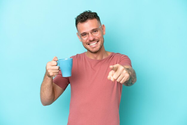 Un joven caucásico sosteniendo una taza de café aislado de fondo azul te señala con una expresión de confianza