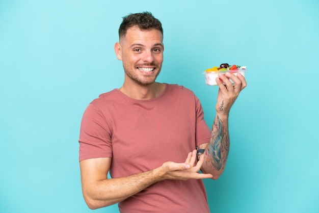 Joven caucásico sosteniendo un tazón de fruta aislado de fondo azul extendiendo las manos a un lado para invitar a venir