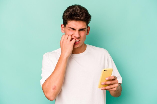 Joven caucásico sosteniendo un teléfono móvil aislado de fondo azul mordiéndose las uñas nervioso y muy ansioso