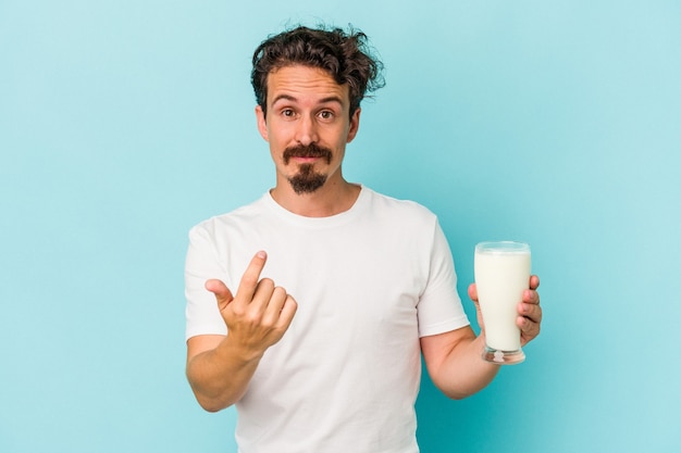 Joven caucásico sosteniendo un vaso de leche aislado sobre fondo azul apuntando con el dedo hacia usted como si estuviera invitando a acercarse.