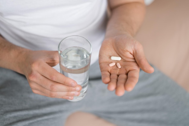 Un joven caucásico sufre de una enfermedad, toma pastillas, sostiene un vaso de agua, se sienta en la cama en la vista superior del dormitorio