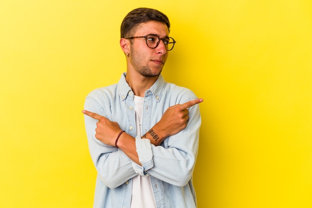 Joven caucásico con tatuajes aislados sobre fondo amarillo apunta hacia los lados, está tratando de elegir entre dos opciones.