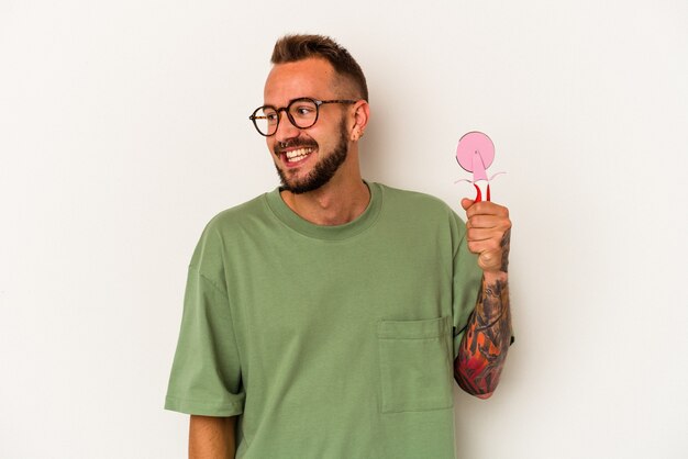 Joven caucásico con tatuajes sosteniendo piruleta aislado sobre fondo blanco mira a un lado sonriente, alegre y agradable.