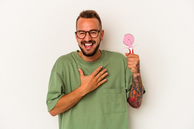 Joven caucásico con tatuajes sosteniendo piruleta aislado sobre fondo blanco se ríe a carcajadas manteniendo la mano en el pecho.