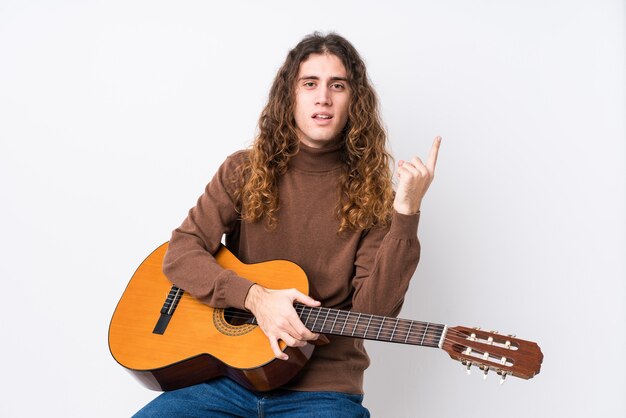 Joven caucásico tocando la guitarra aislado sonriendo y apuntando a un lado, mostrando algo en el espacio en blanco.