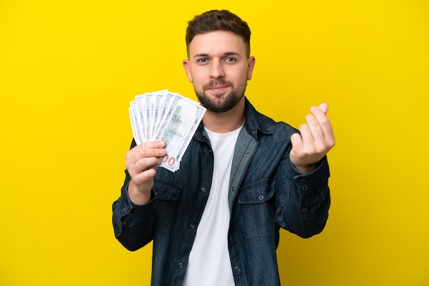 Joven caucásico tomando mucho dinero aislado de fondo amarillo haciendo gestos de dinero