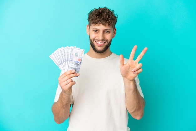 Joven caucásico tomando mucho dinero aislado de fondo azul feliz y contando tres con los dedos