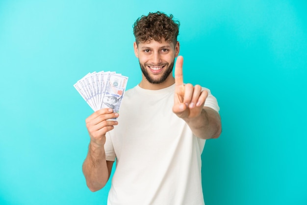 Joven caucásico tomando mucho dinero aislado de fondo azul mostrando y levantando un dedo