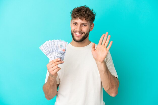 Joven caucásico tomando mucho dinero aislado de fondo azul saludando con la mano con expresión feliz