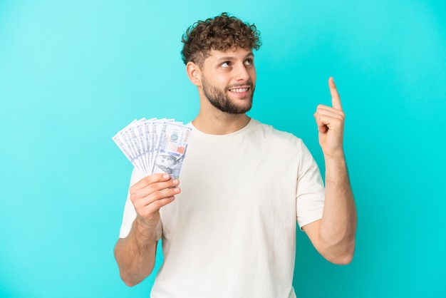 Joven caucásico tomando mucho dinero aislado de fondo azul señalando una gran idea