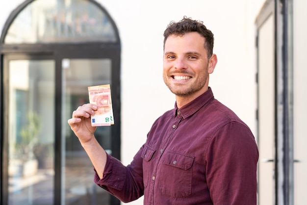 Foto joven caucásico tomando mucho dinero al aire libre sonriendo mucho