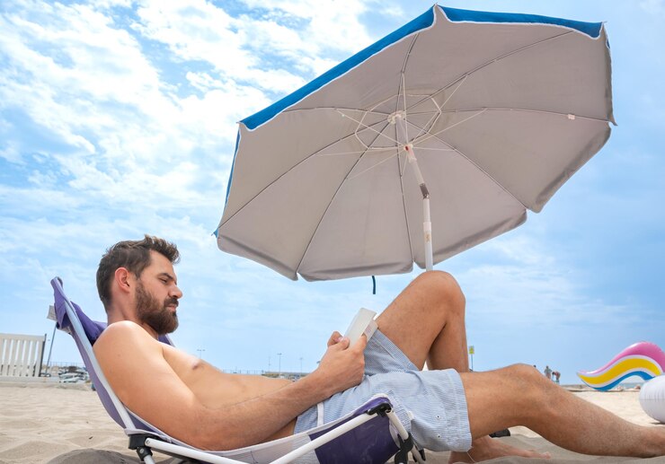 Joven centrado en la lectura tendido en la playa con paraguas y flotador inflable