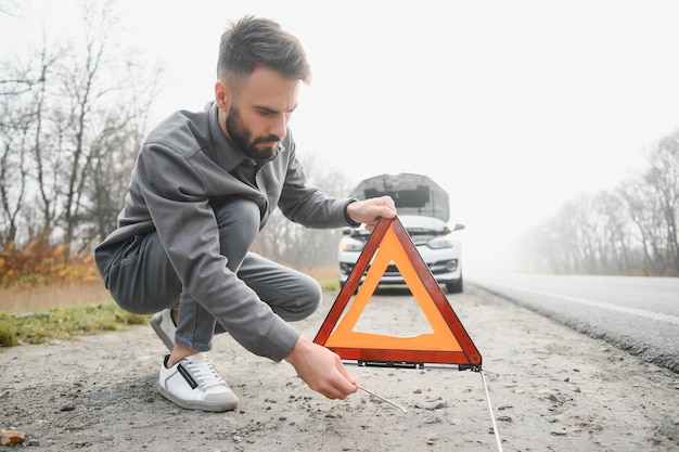 Un joven cerca de un auto roto con el capó abierto en la carretera