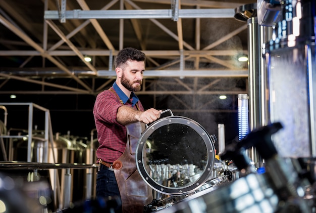 Joven cervecero en delantal de cuero que supervisa el proceso de fermentación de la cerveza en la fábrica de cerveza moderna ...