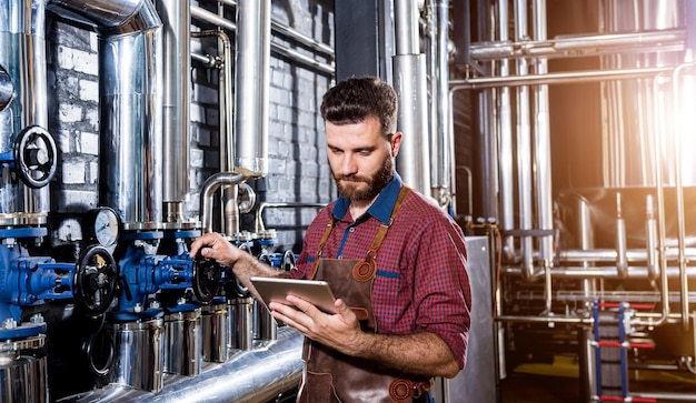 Joven cervecero en delantal de cuero que supervisa el proceso de fermentación de la cerveza en la fábrica de cerveza moderna ...