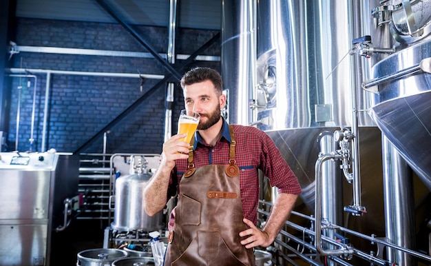 Joven cervecero vistiendo un delantal de cuero está probando cerveza en una cervecería moderna