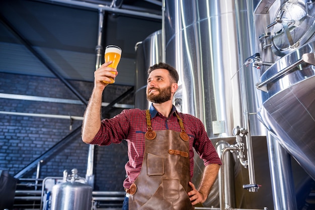 Foto joven cervecero vistiendo un delantal de cuero está probando cerveza en una cervecería moderna
