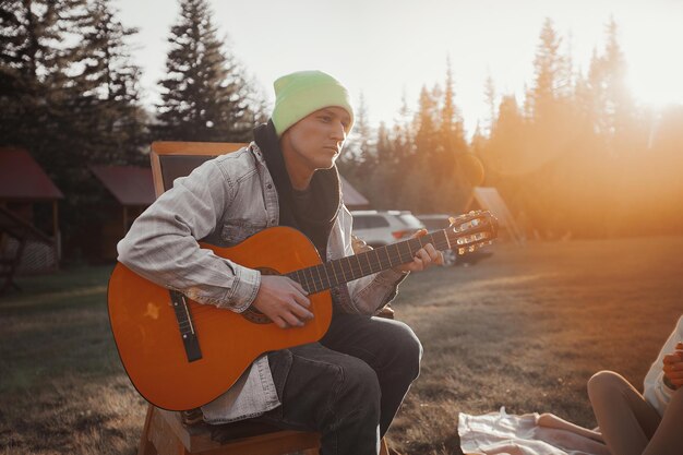 Un joven con una chaqueta vaquera y un sombrero está sentado y tocando una guitarra acústica en el fondo de un centro recreativo y puesta de sol. Pasar tiempo en el campamento.