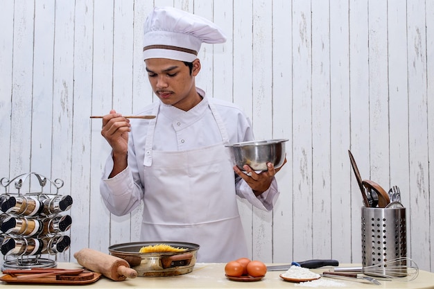 Joven chef asiático con toque blanco y delantal con tazón y espátula de madera probando la comida