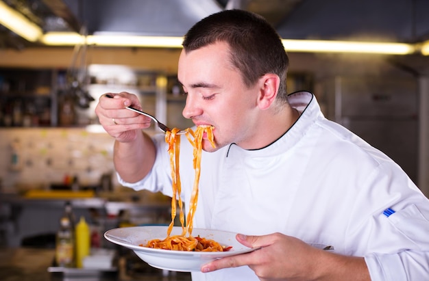Joven chef atractivo sosteniendo un plato y probando la pasta. Chef cocinero degustación de comida con tenedor en la cocina del restaurante.