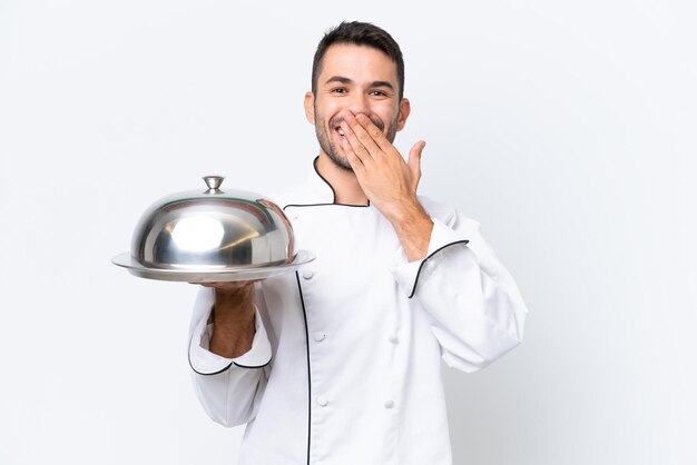 Joven chef con bandeja aislado sobre fondo blanco feliz y sonriente cubriendo la boca con la mano