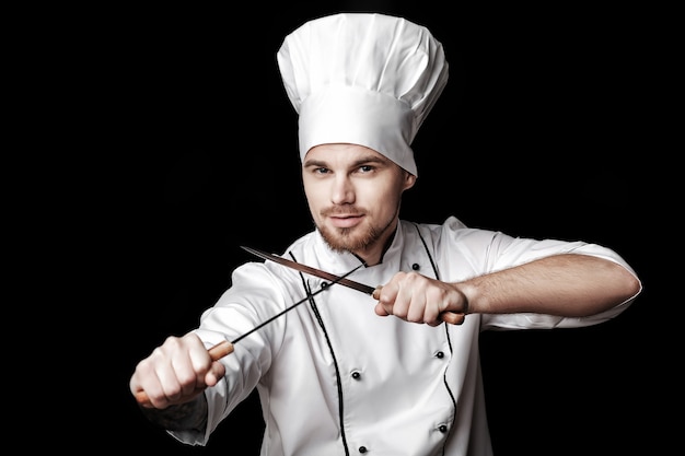 Joven chef barbudo en uniforme blanco sosteniendo dos cuchillos sobre fondo negro