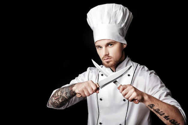 Joven chef barbudo en uniforme blanco tiene dos cuchillos sobre fondo negro