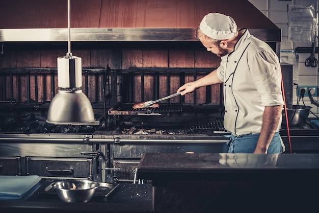 Joven chef blanco en delantal azul y sombrero de pie cerca del brasero con brasas hombre cocinando filete de ternera