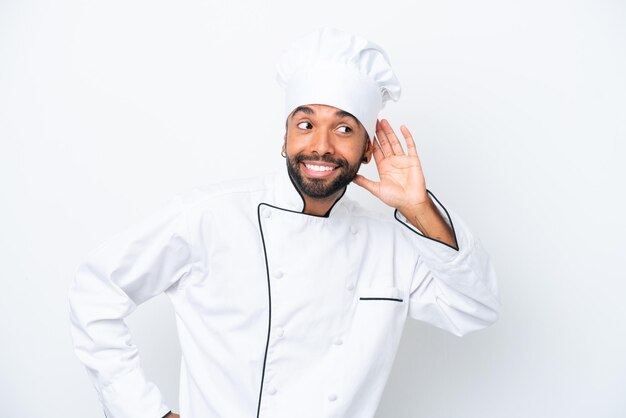 Joven chef brasileño aislado de fondo blanco escuchando algo poniendo la mano en la oreja