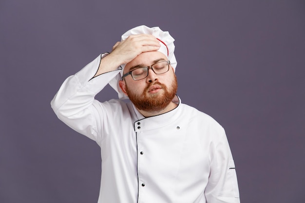 Joven chef cansado con uniforme de anteojos y gorra manteniendo la mano en la cabeza con los ojos cerrados aislados en el fondo morado