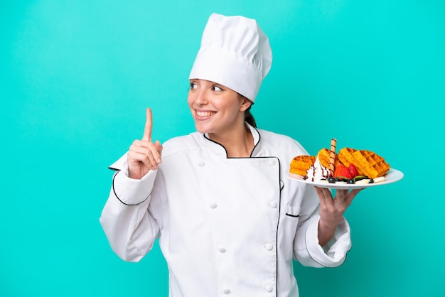 Foto joven chef caucásica sosteniendo gofres aislados de fondo azul con la intención de darse cuenta de la solución mientras levanta un dedo