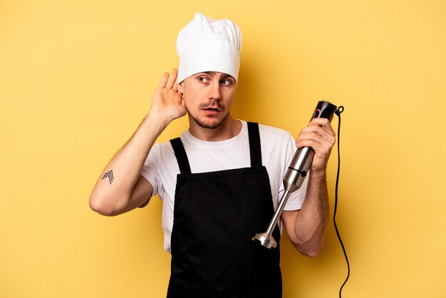 Joven chef caucásico sosteniendo una licuadora aislada en un fondo amarillo tratando de escuchar un chisme.