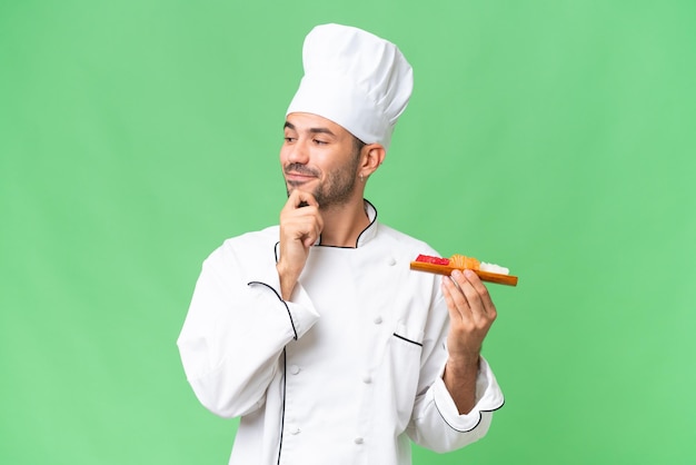 Joven chef caucásico sosteniendo un sushi sobre un fondo aislado mirando hacia un lado y sonriendo