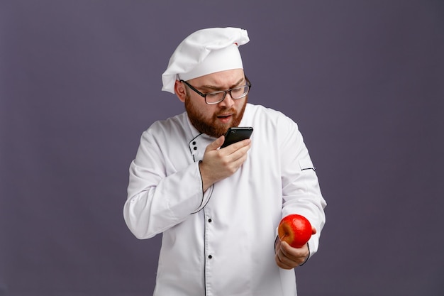 Joven chef concentrado con uniforme de anteojos y gorra sosteniendo una manzana tomándole una foto con un teléfono móvil aislado en un fondo morado