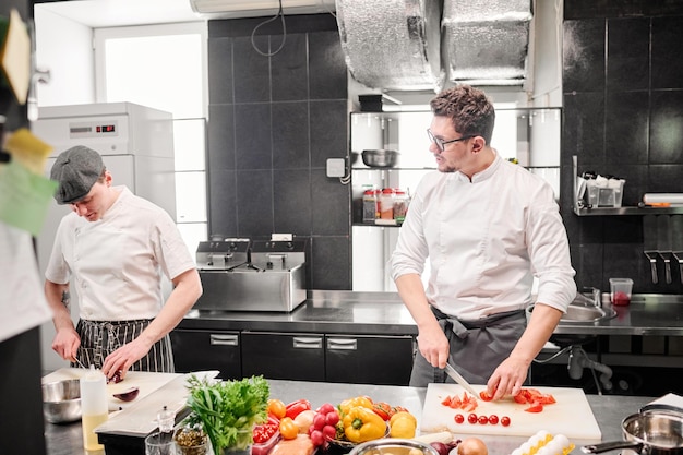 Joven chef cortando tomates en la tabla de cortar y discutiendo el menú con su asistente durante su trabajo en la mesa de la cocina