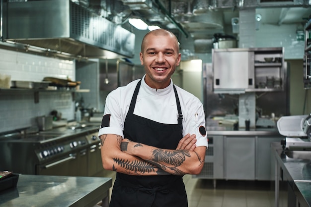 Joven chef en delantal manteniendo los brazos tatuados cruzados y sonriendo mientras está de pie en la cocina de un restaurante