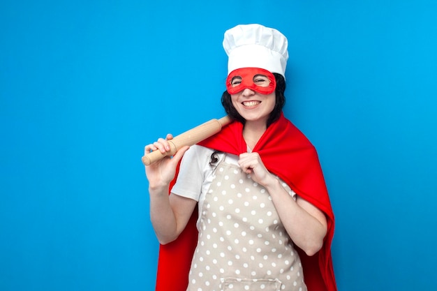 Una joven chef en disfraz de superhombre sostiene un artículo de cocina en un fondo azul mujer ama de casa