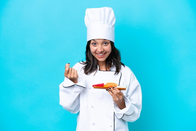 Joven chef hispana mujer sosteniendo sashimi aislado sobre fondo azul haciendo gesto de dinero