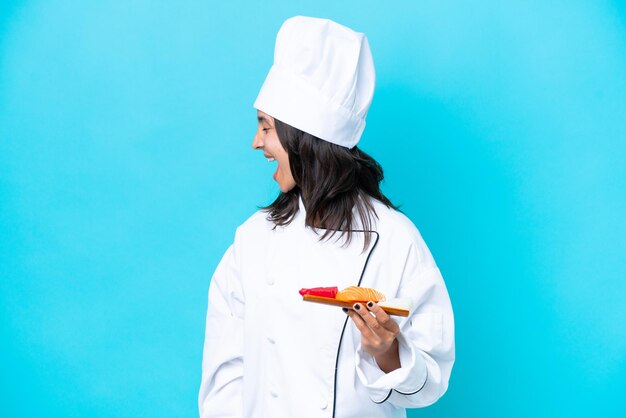 Joven chef hispana mujer sosteniendo sashimi aislado sobre fondo azul riendo en posición lateral