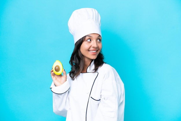 Joven chef hispana sosteniendo aguacate aislado de fondo azul pensando en una idea mientras mira hacia arriba