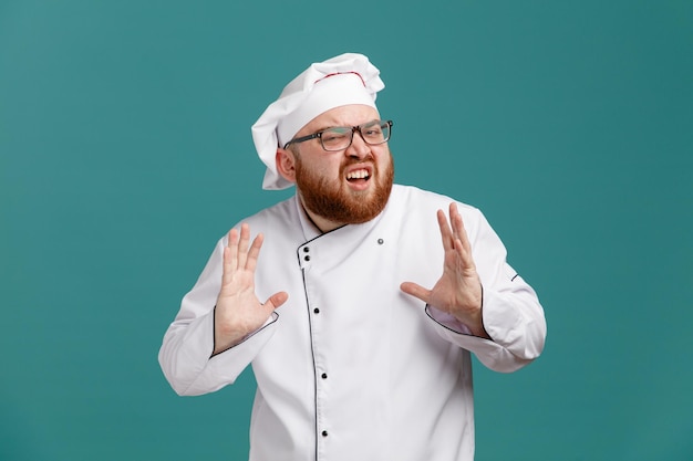 Un joven chef irritado con uniforme de gafas y gorra mirando a la cámara mostrando las manos vacías aisladas de fondo azul