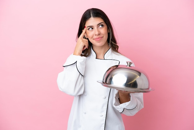 Joven chef italiana sosteniendo una bandeja con tapa aislada en un fondo rosa con dudas y pensando