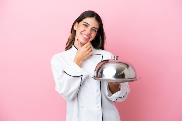 Joven chef italiana sosteniendo una bandeja con tapa aislada de fondo rosa feliz y sonriente