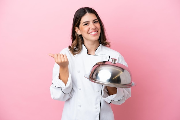 Joven chef italiana sosteniendo una bandeja con tapa aislada en un fondo rosa haciendo gestos de dinero