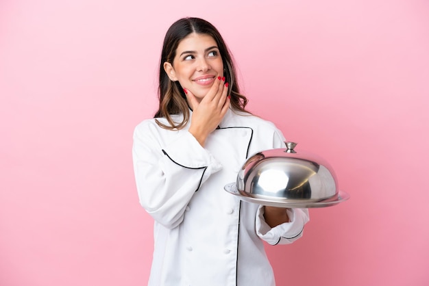 Joven chef italiana sosteniendo una bandeja con tapa aislada en un fondo rosa mirando hacia arriba mientras sonríe