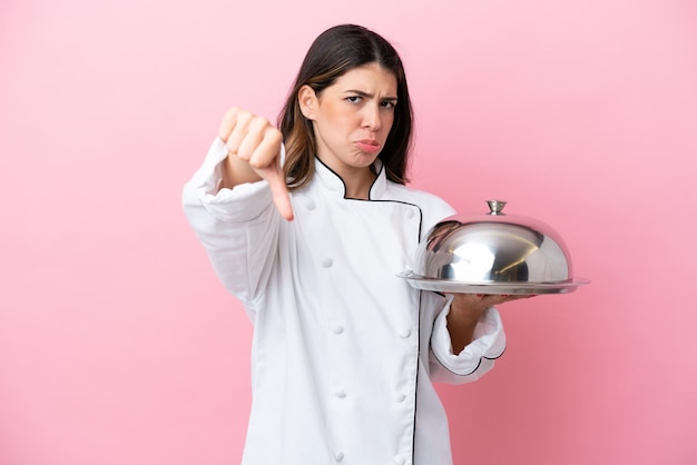 Joven chef italiana sosteniendo una bandeja con tapa aislada en un fondo rosa mostrando el pulgar hacia abajo con expresión negativa