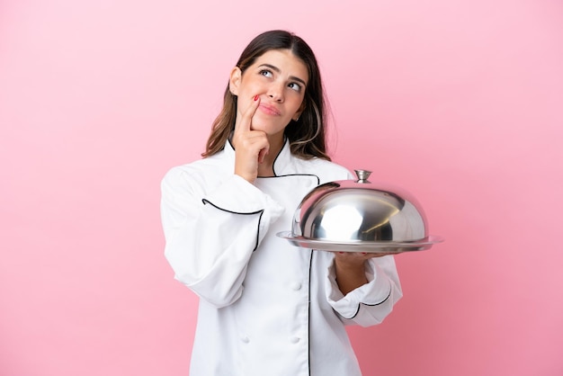 Joven chef italiana sosteniendo una bandeja con tapa aislada en un fondo rosado que tiene dudas mientras mira hacia arriba